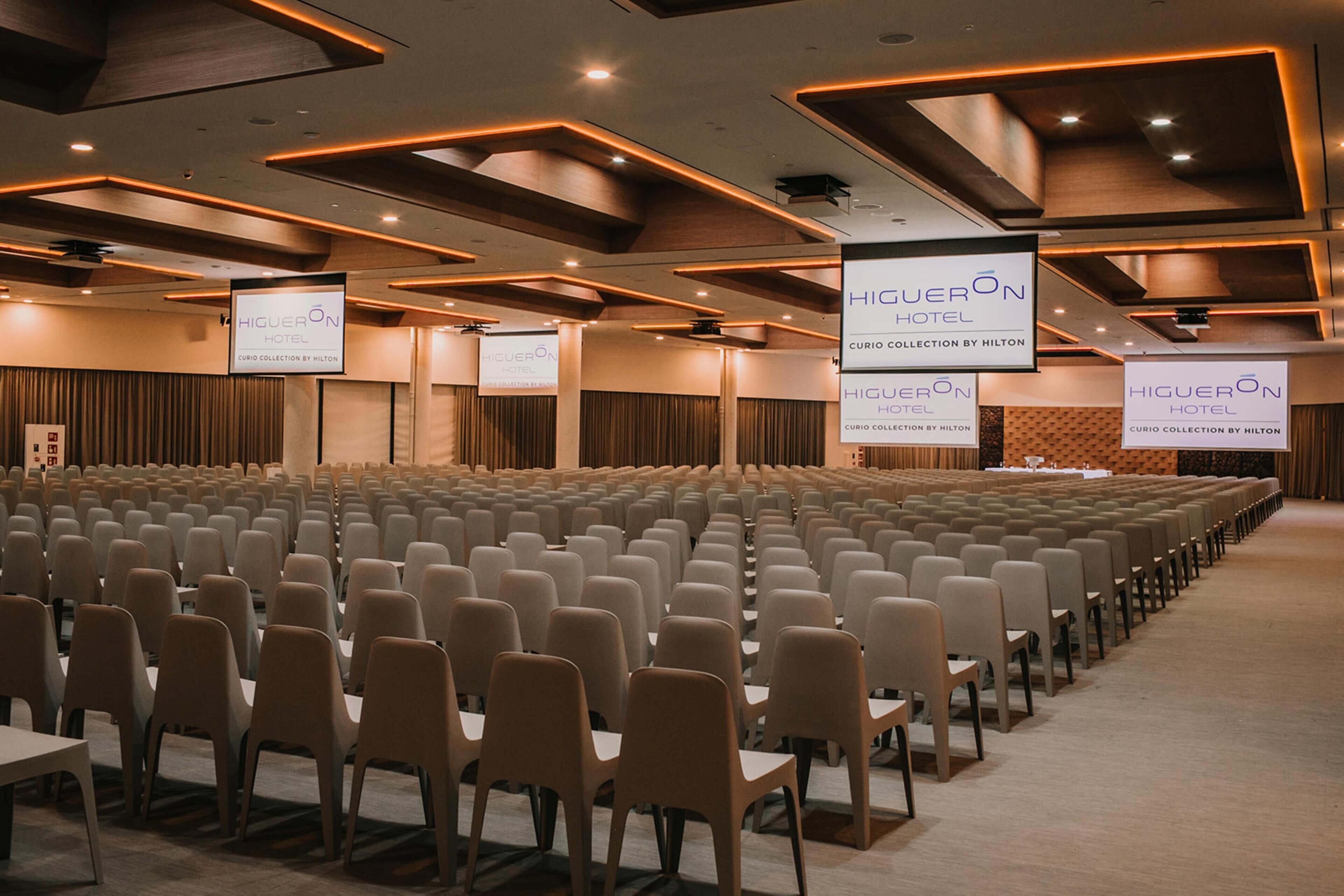 Un salón de conferencias con filas de sillas y un escenario con una pantalla que muestra 'HIGUERON HOTEL' y 'CURIO COLLECTION BY HILTON'.