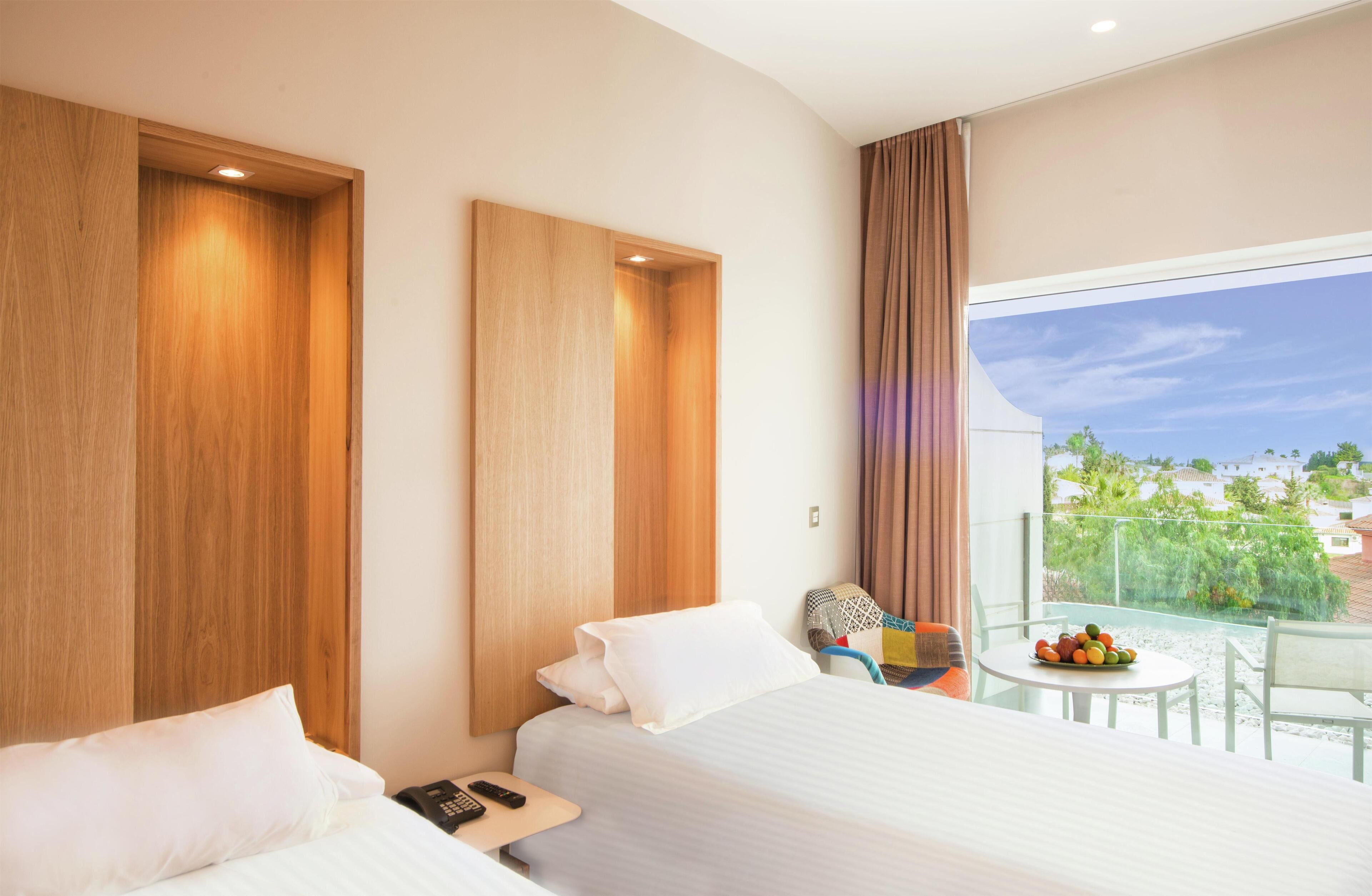 A hotel room with a large bed, a small table with fruit, a chair, and a balcony with a view.