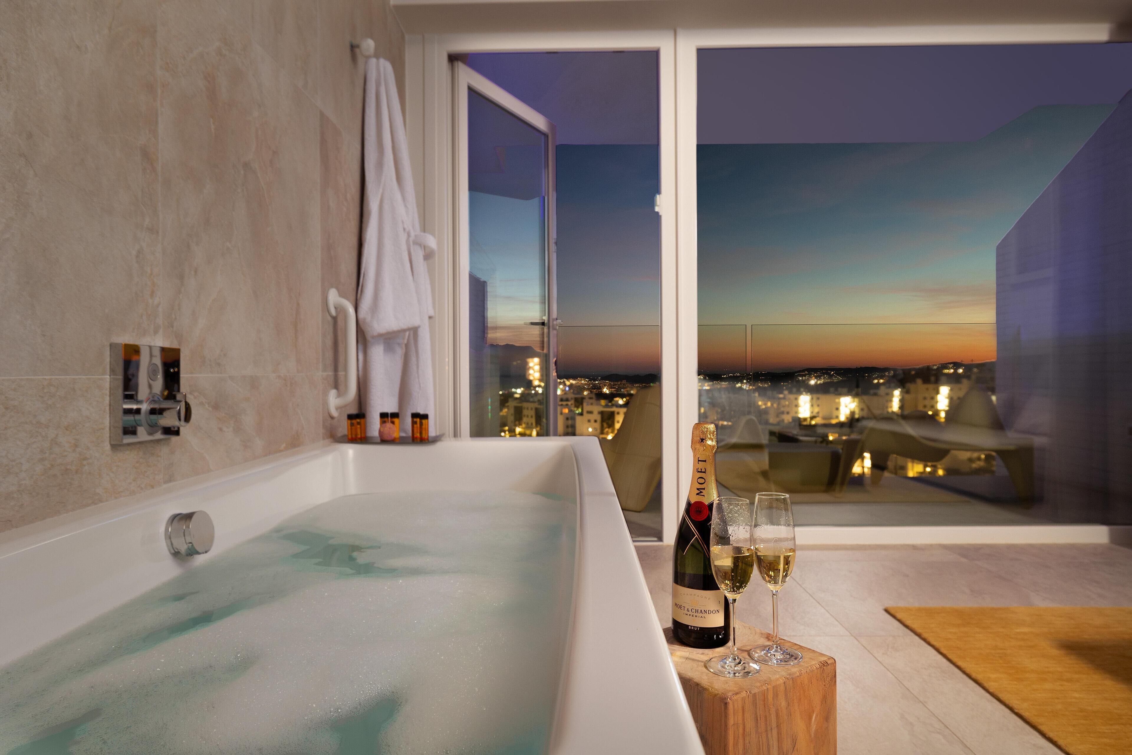 A modern bathroom with a large bathtub, a bottle of champagne, and a glass of champagne on a wooden side table.