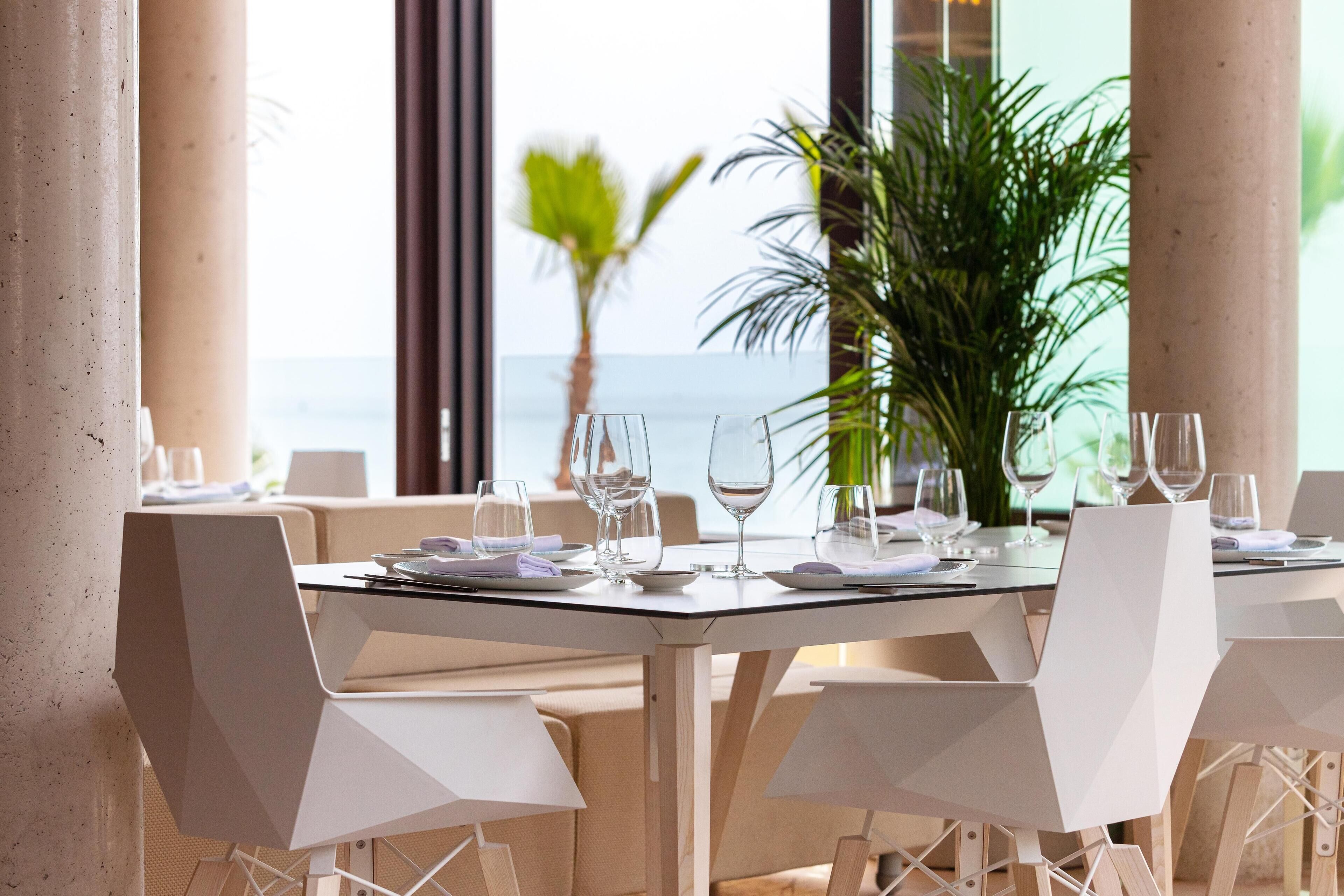 Comedor con vistas al mar, con una mesa dispuesta con vasos y platos, rodeada de sillas blancas.