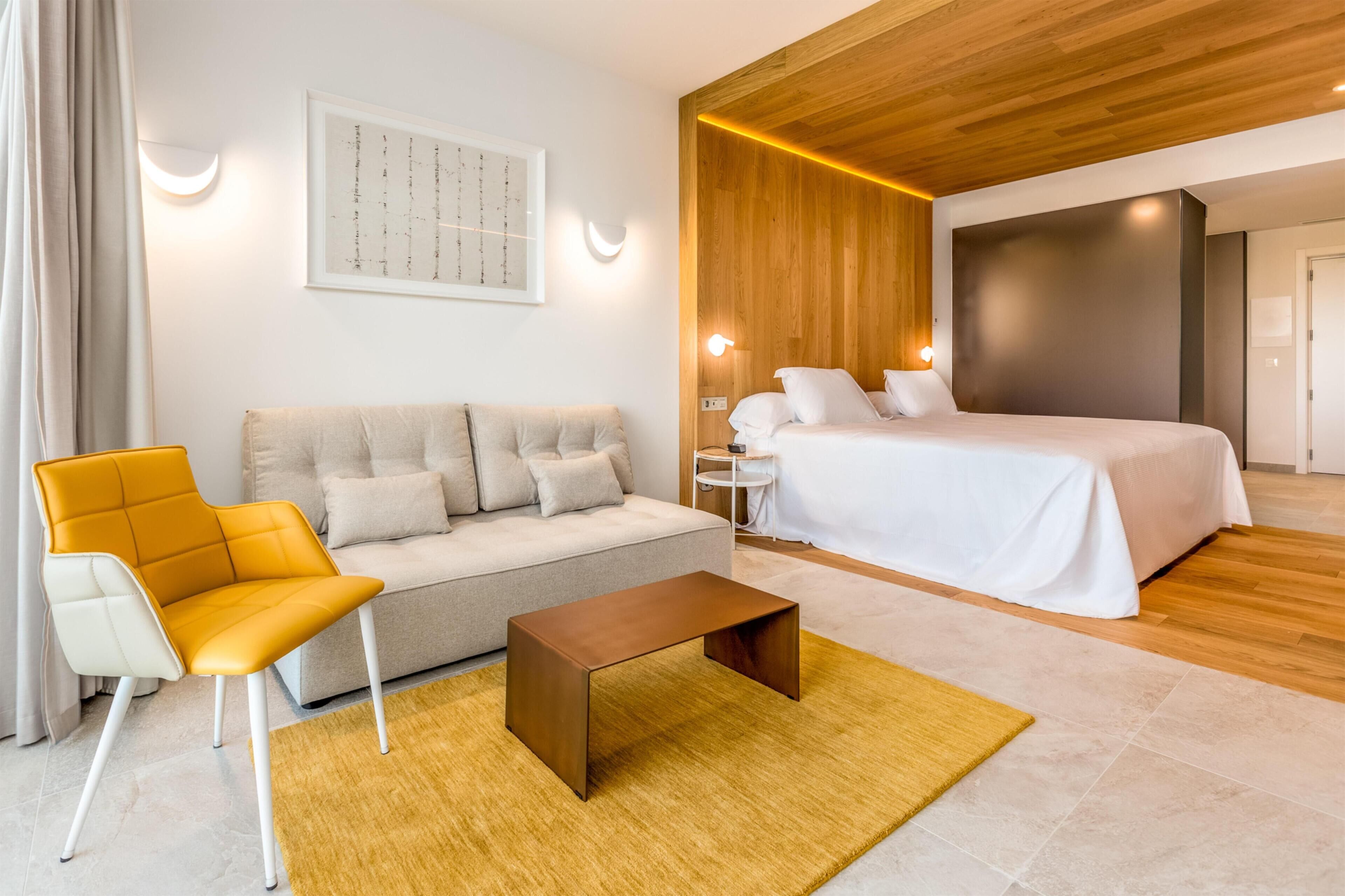 A modern hotel room with a wooden ceiling, a bed, a sofa, a coffee table, and a wall-mounted artwork.