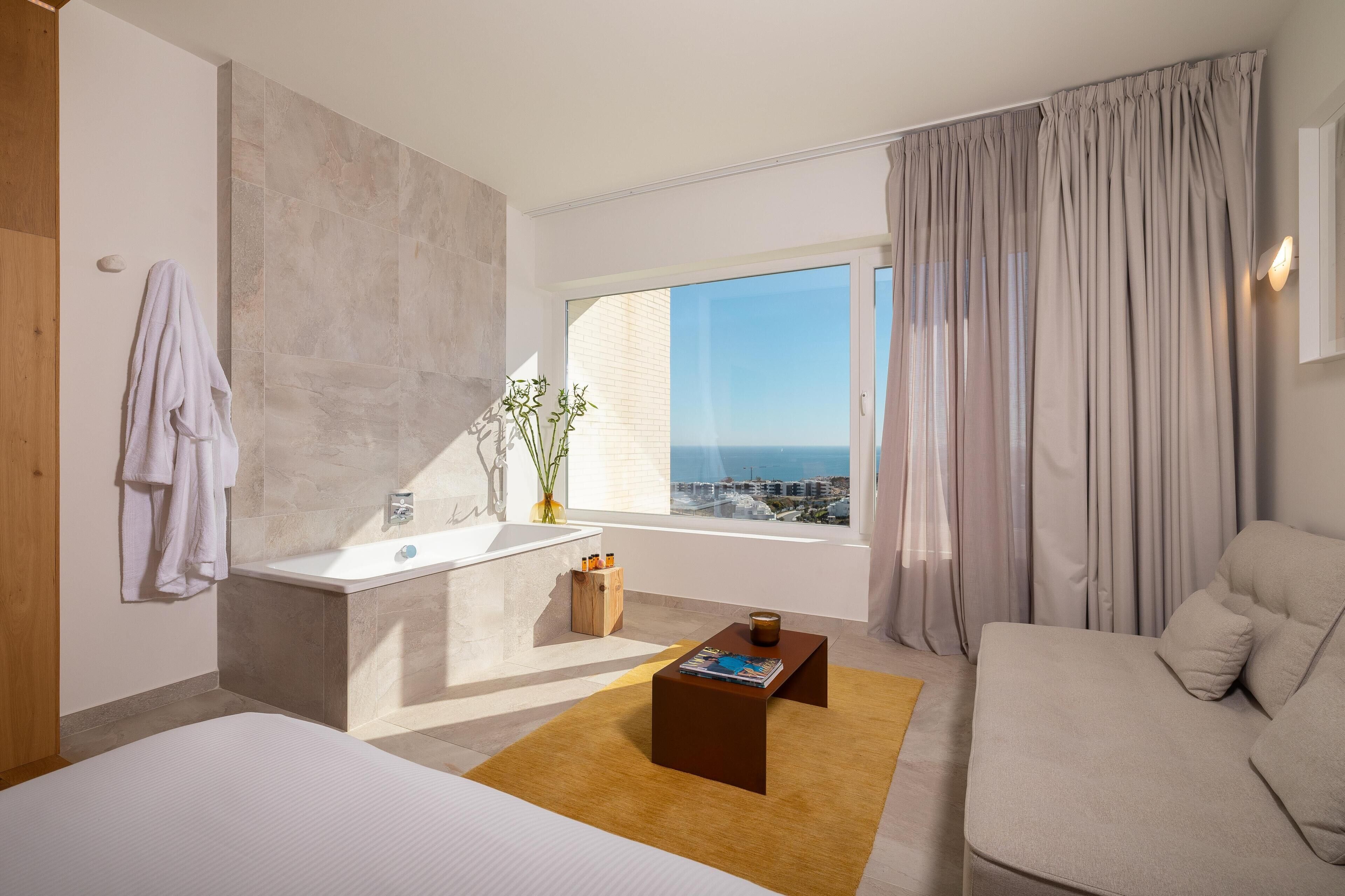 Una habitación de hotel moderna con un ventanal grande, una bañera, un sofá y una mesa de centro.