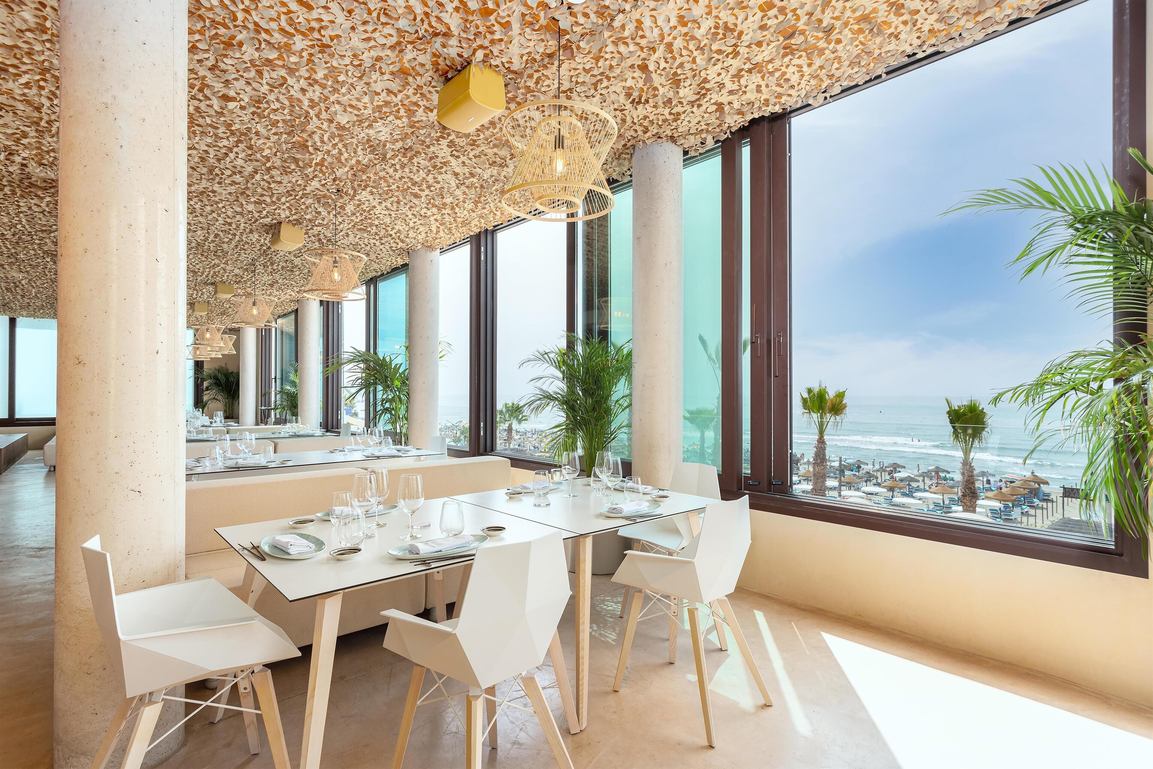 A dining area with a view of the beach, featuring a table set with white chairs and a glass-top table.