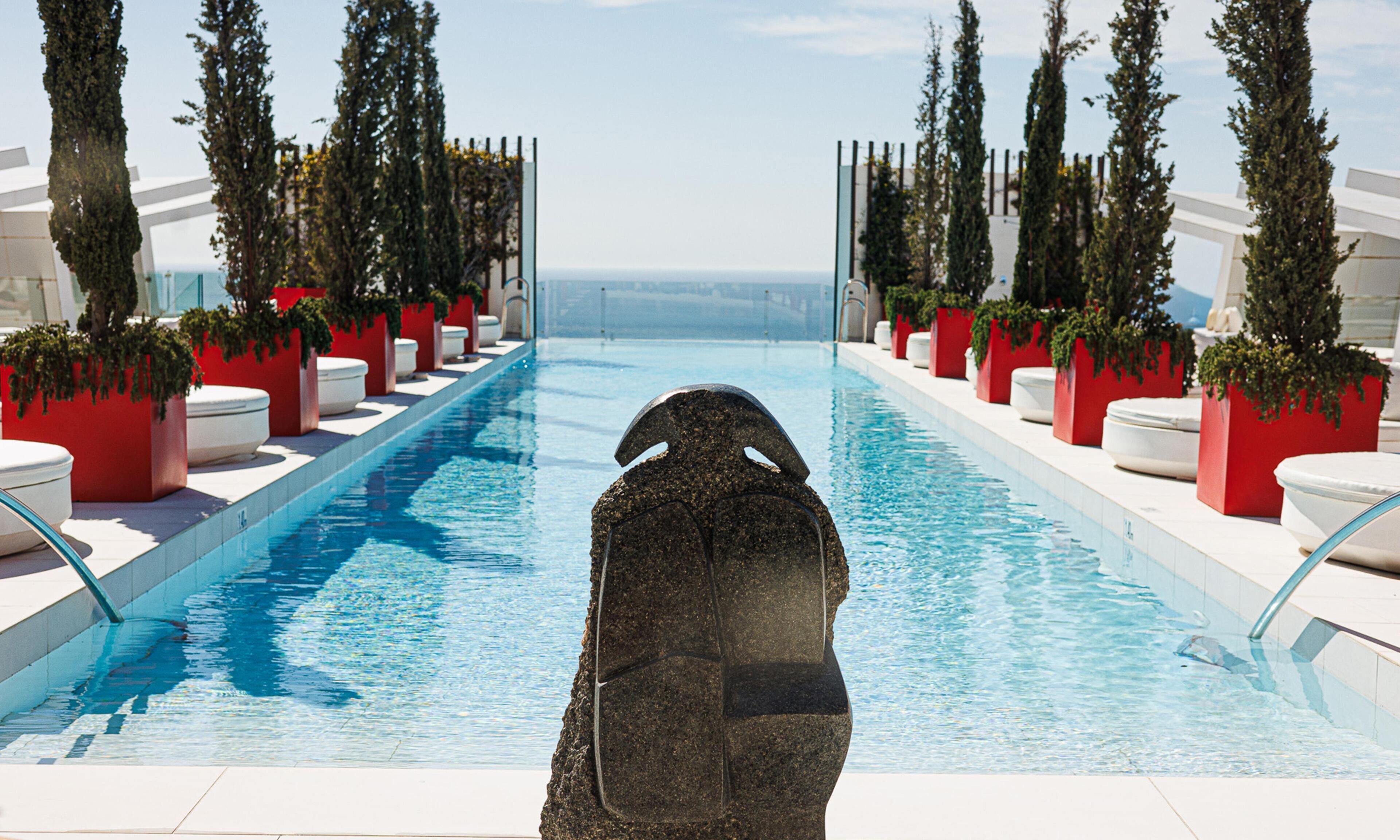 A pool with a stone sculpture in the center, surrounded by red planters and white lounge chairs.