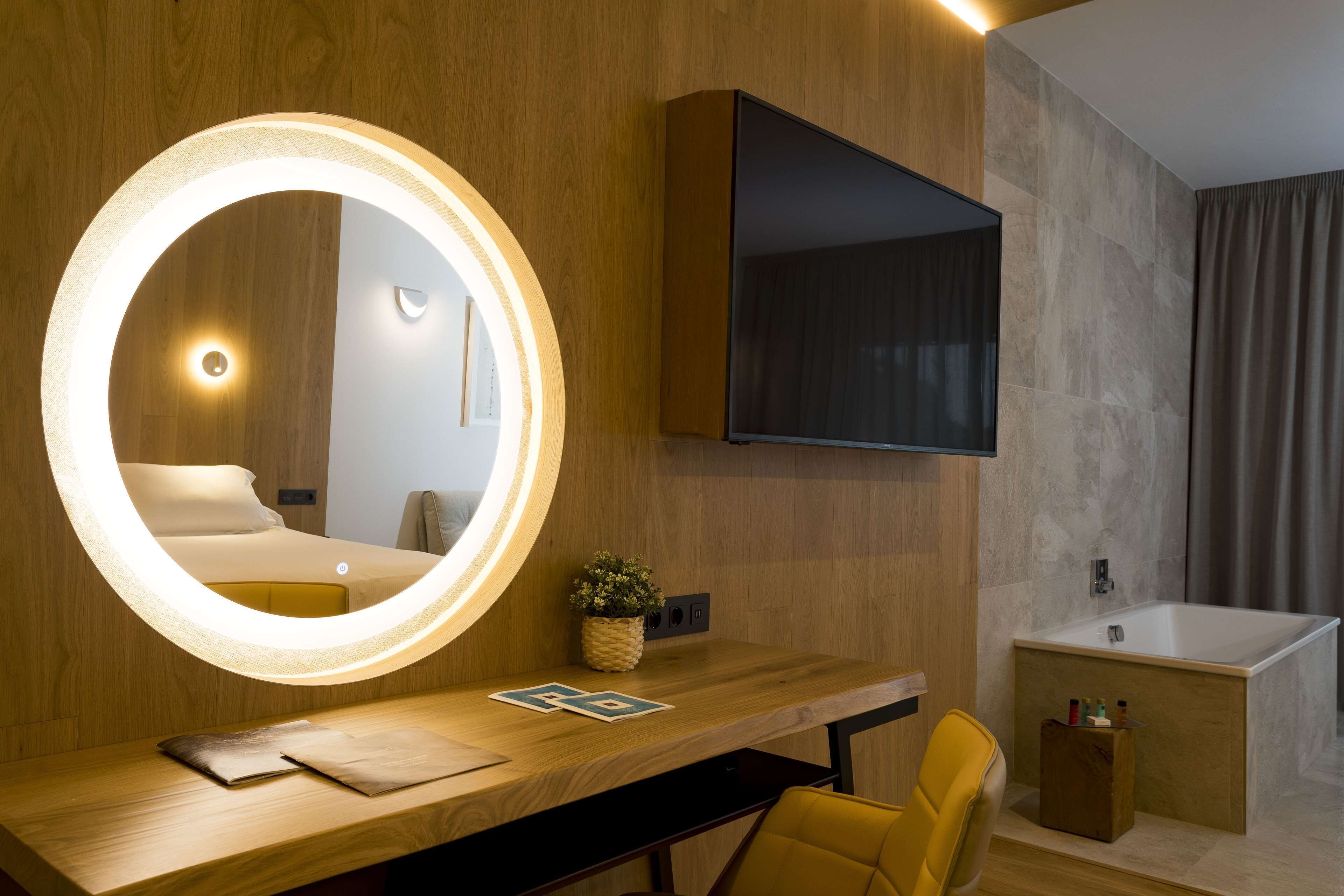 Una habitación de hotel moderna con un escritorio de madera, un espejo redondo con iluminación, un televisor de pantalla plana, una bañera y una silla amarilla.