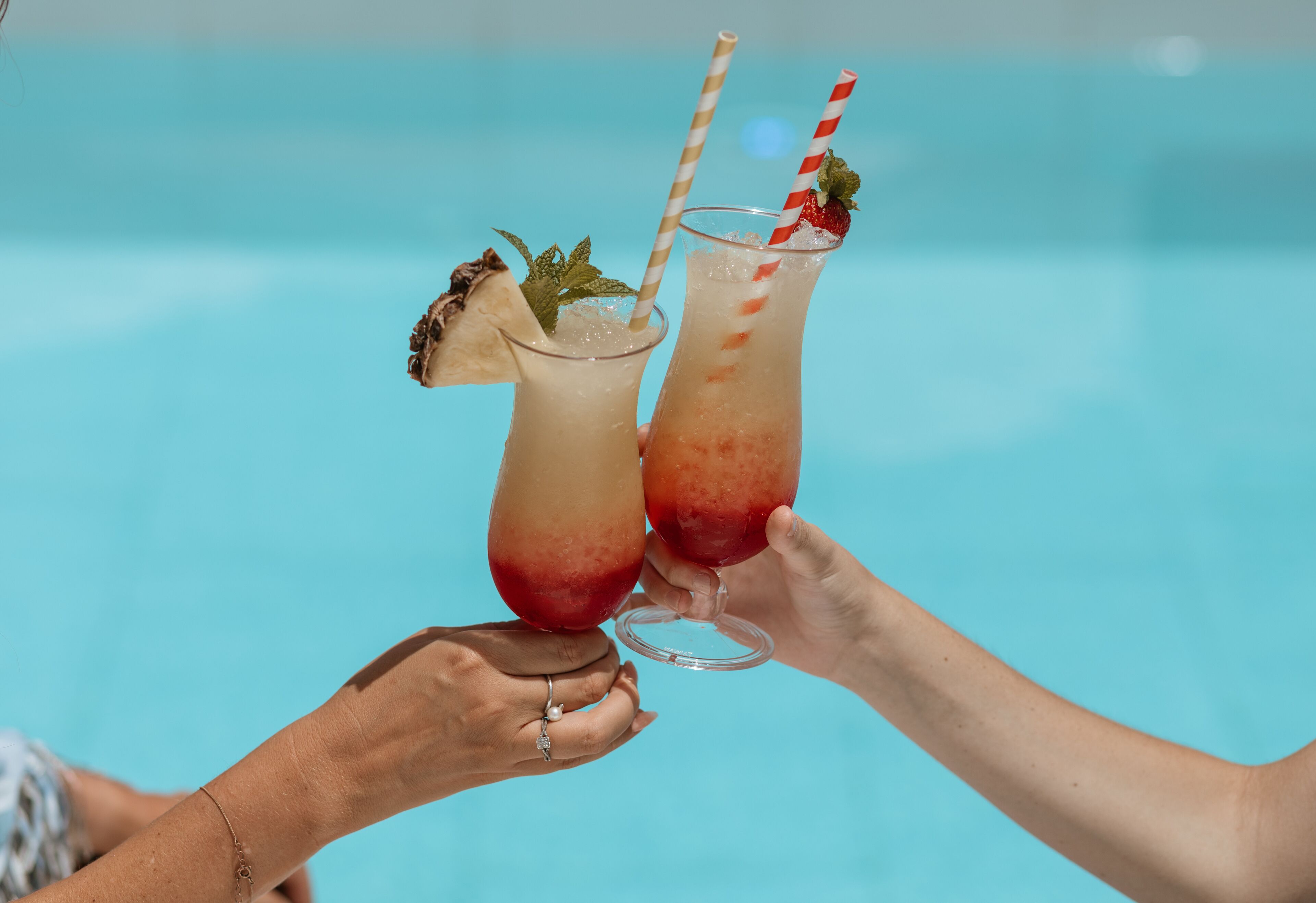 Two people holding cocktail glasses with straws and garnishes by a pool.