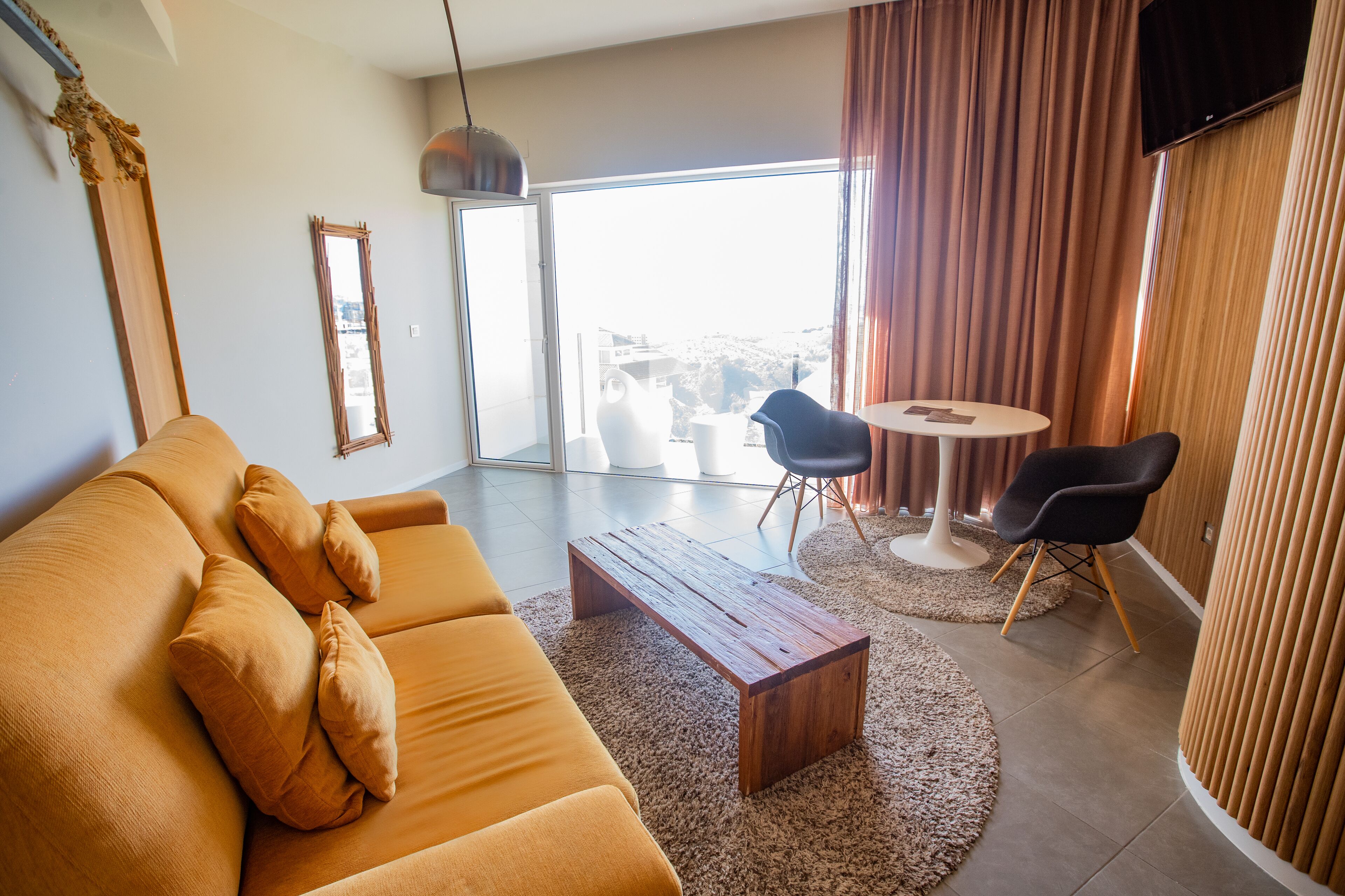 Un salon moderne comprenant un canapé, une table basse et des fauteuils, une grande fenêtre avec des rideaux et un téléviseur à écran plat fixé au mur.