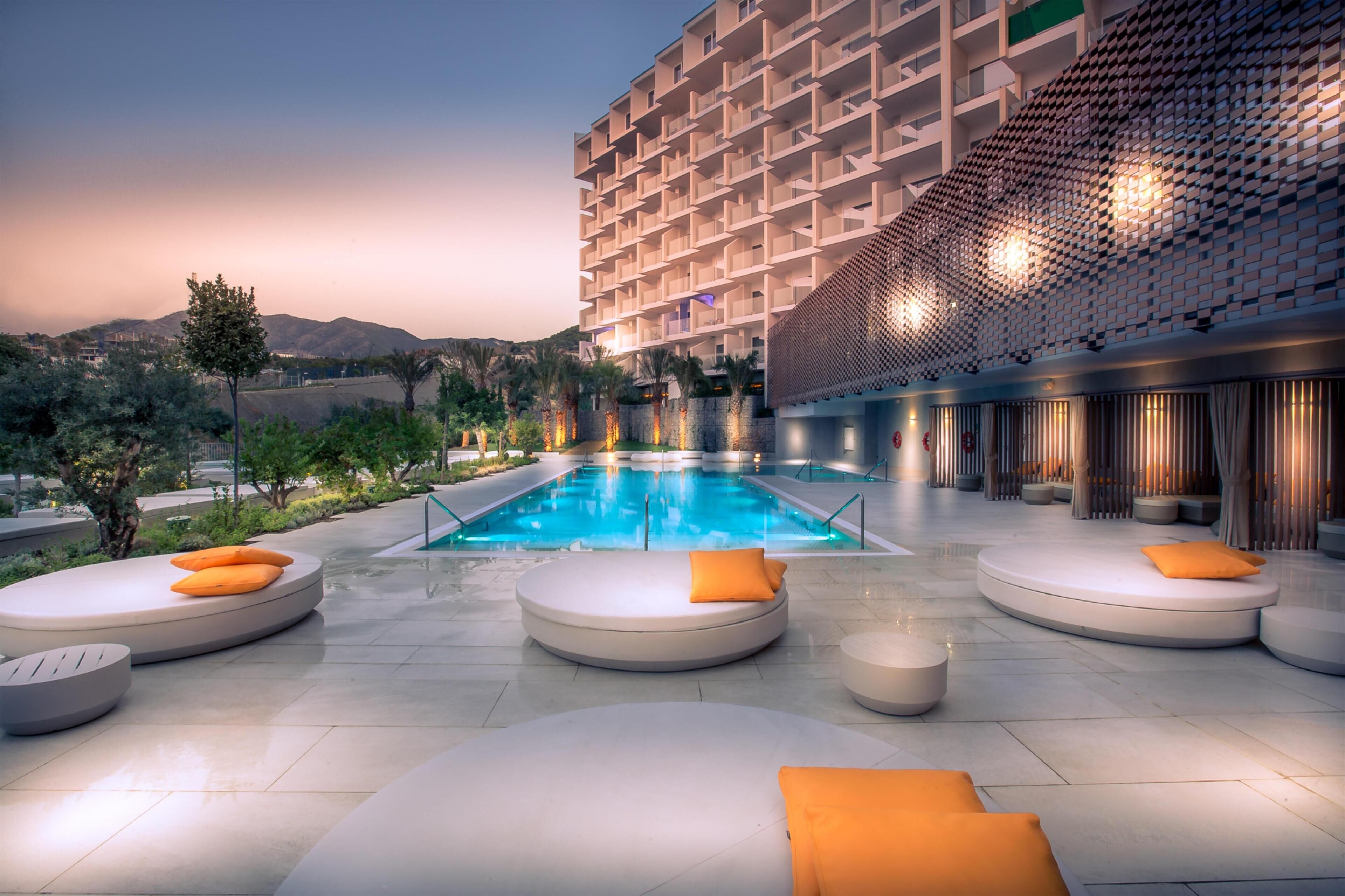 A modern hotel with a rooftop pool, outdoor seating, and a view of the mountains.
