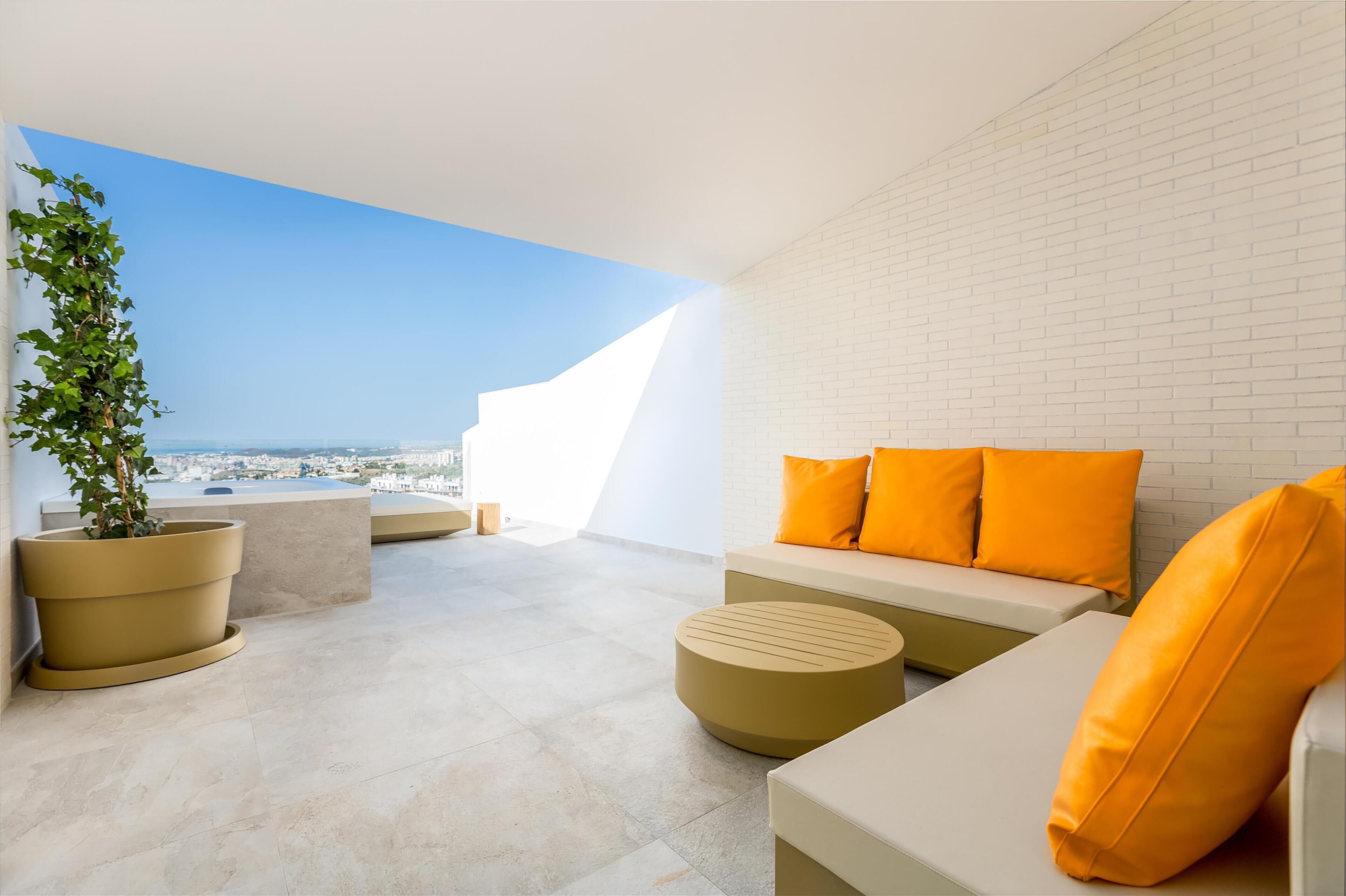A modern outdoor seating area with a white brick wall, a large potted plant, and orange cushions.