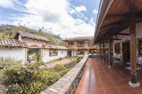 A courtyard with a tiled floor, a covered seating area with tables and chairs, and a garden with various plants and flowers.