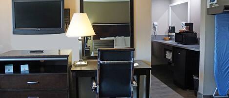 A hotel room with a desk, chair, TV, lamp, and a kitchenette area.
