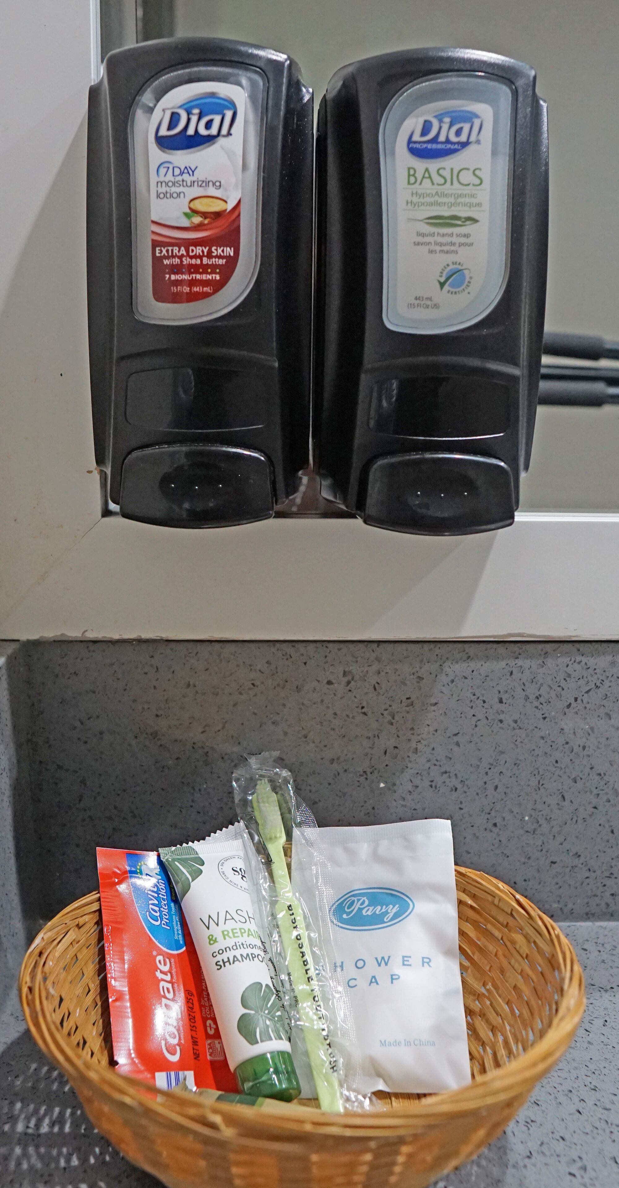 A bathroom with a hand soap dispenser and a hand soap bottle. Below, a basket contains a toothbrush, a bar of soap, a packet of shampoo, and a shower cap.
