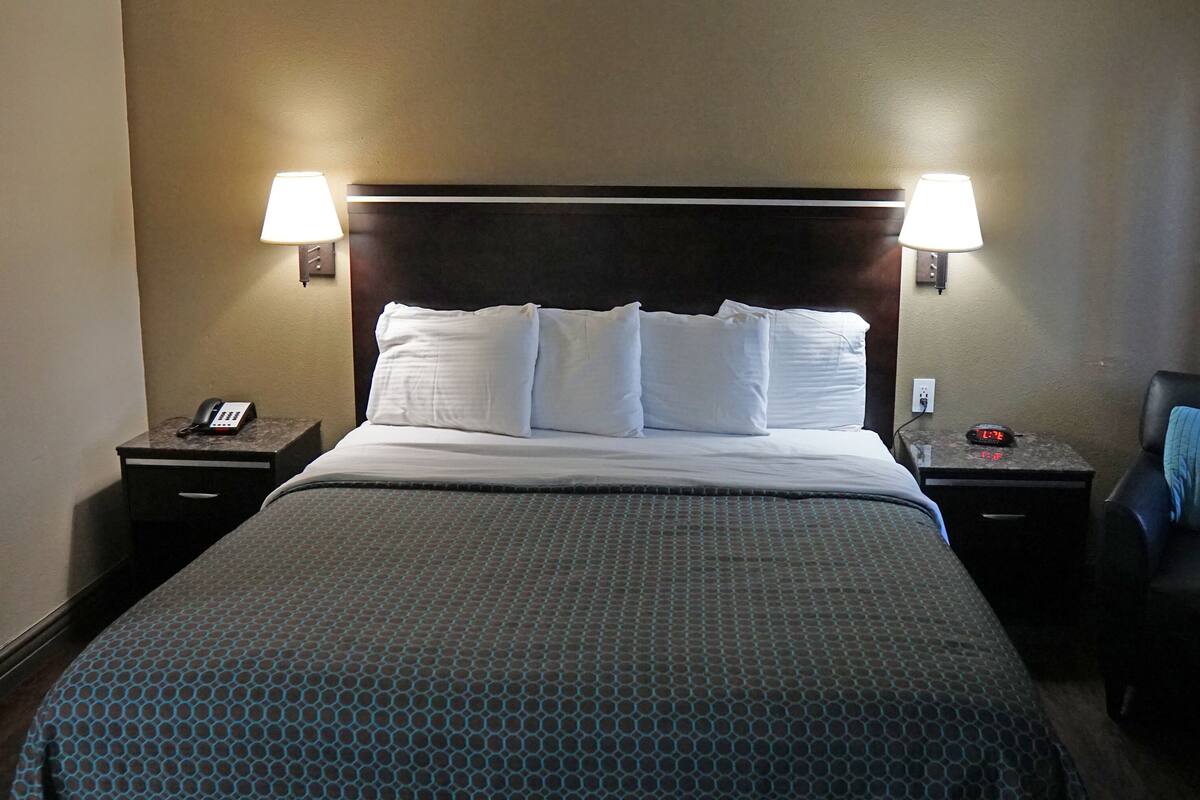 Una habitación de hotel con una cama, dos mesitas de noche, una silla y dos lámparas de pared.