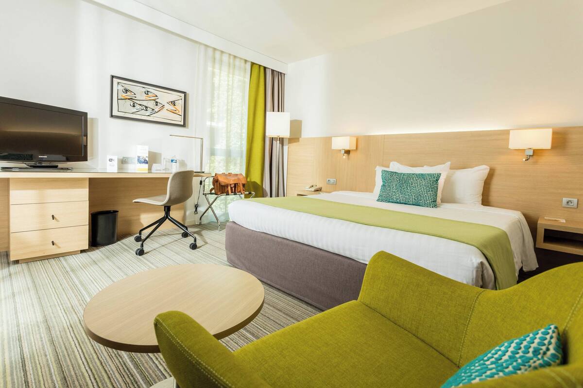 Ein Hotelzimmer mit einem Bett, einem Schreibtisch mit Fernseher, einem Sessel, einem kleinen Tisch und einer Lampe.