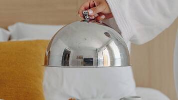 Uma pessoa vestindo um robe branco coloca uma cúpula prateada sobre uma bandeja de café da manhã em uma cama com roupa de cama branca e um travesseiro amarelo.