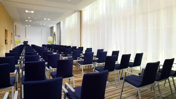A conference room with rows of blue chairs, a podium, and a large window with sheer curtains.