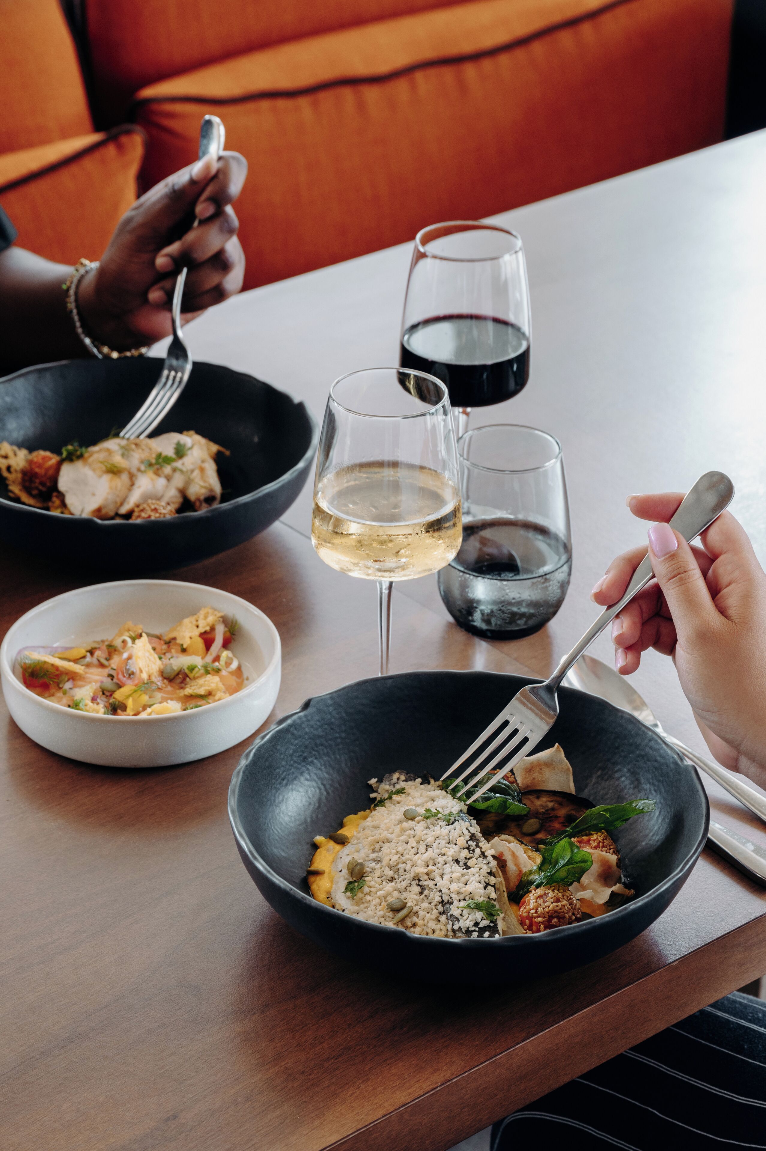 A dining table with three bowls of food, two glasses of wine, and a person holding a fork.