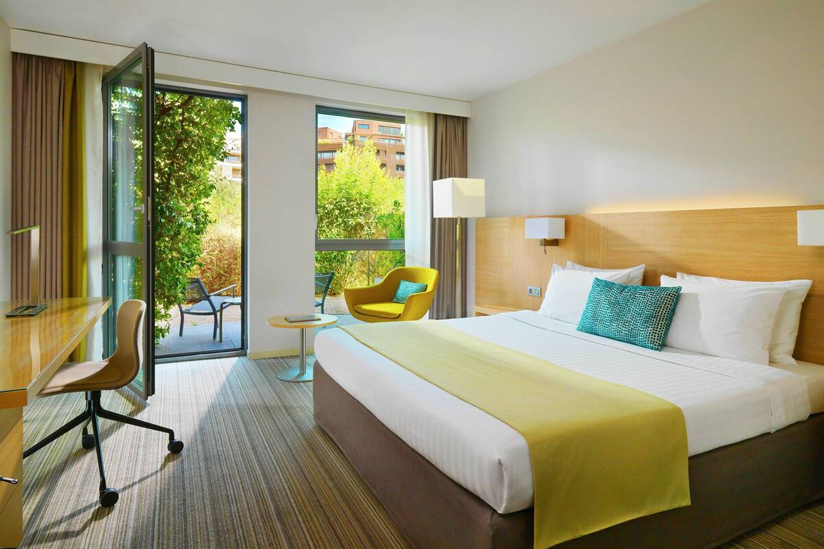 Ein Hotelzimmer mit einem großen Bett, einem Schreibtisch, einem Stuhl und Blick ins Freie.