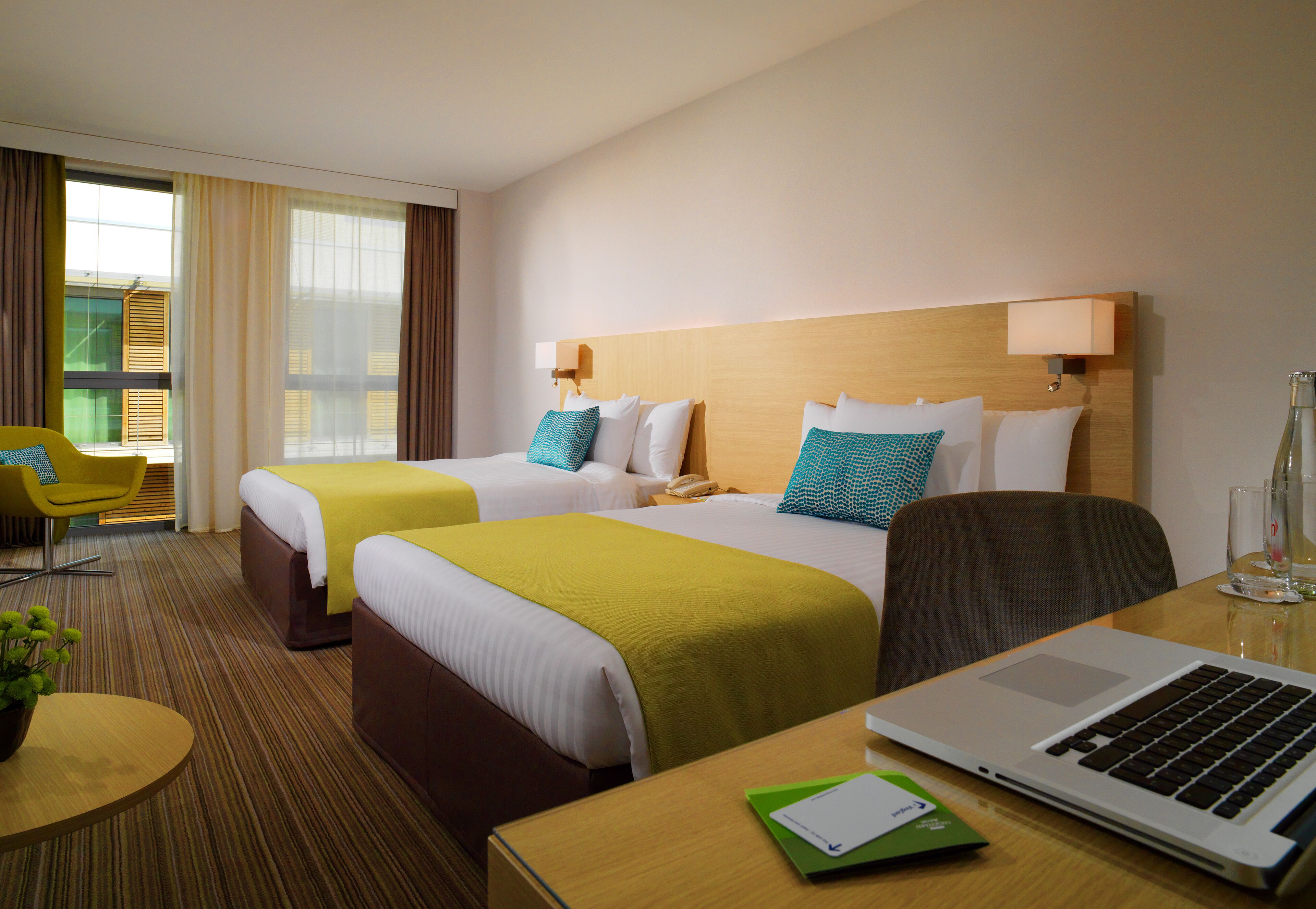 A hotel room with two beds, a desk with a laptop, a chair, and a small table with a plant.