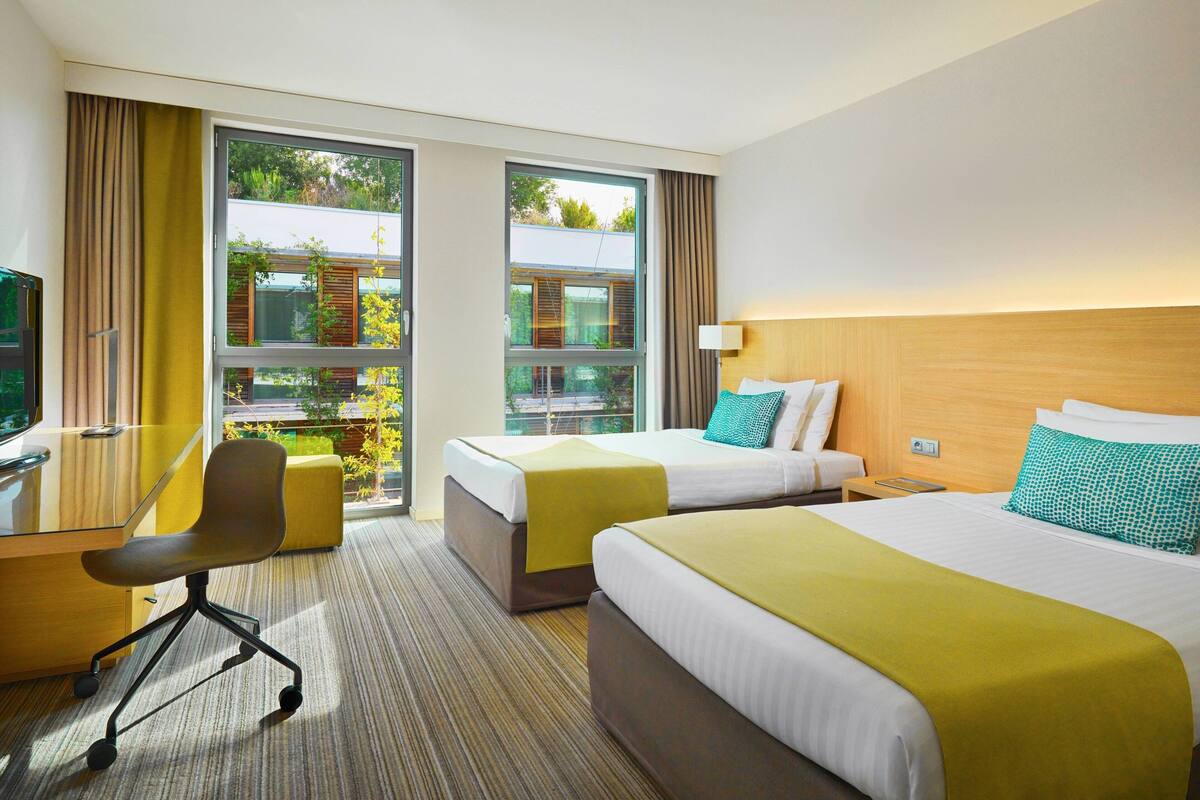 Ein Hotelzimmer mit zwei Betten, einem Schreibtisch, einem Stuhl und großen Fenstern mit Blick auf Grünflächen.