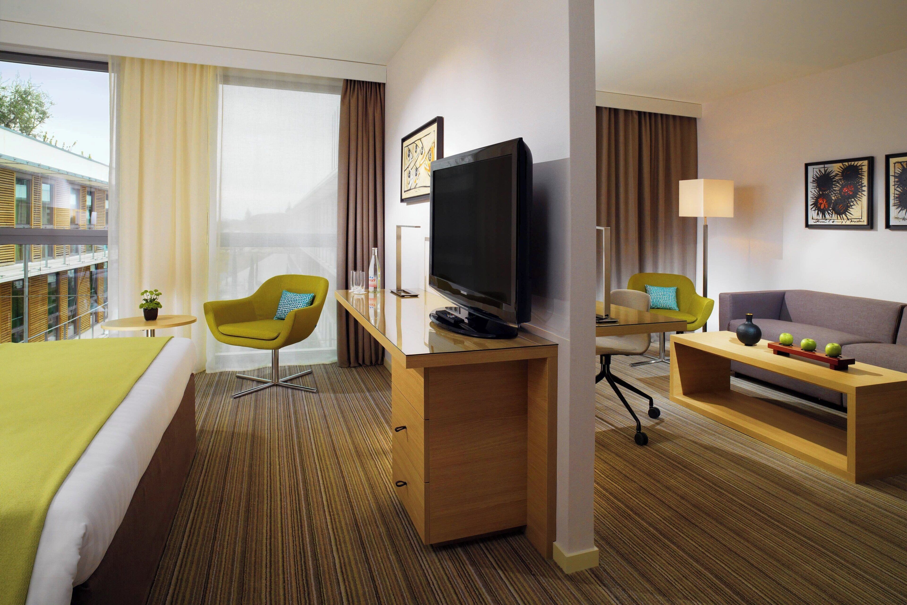 A hotel room with a bed, a desk with a TV, a sofa, and a window with curtains.