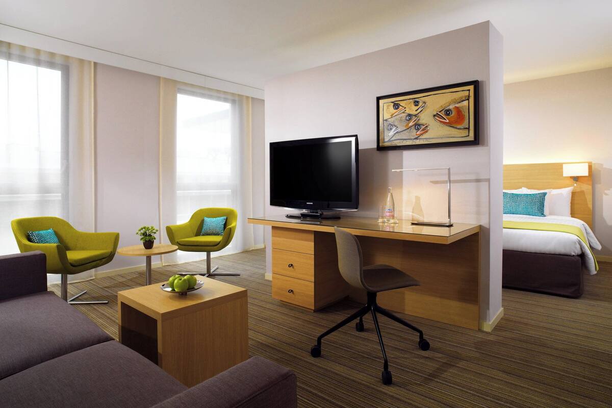 Ein Hotelzimmer mit einem Bett, einem Schreibtisch mit Fernseher, zwei Sesseln, einem Couchtisch und einem Bild an der Wand.