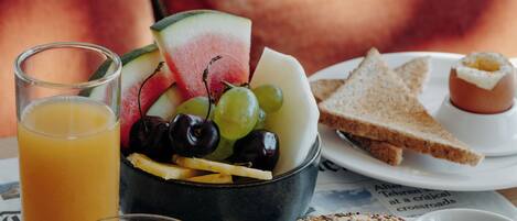 A breakfast setting with a glass of orange juice, a bowl of fruit, croissants, and toast on a table with a newspaper.