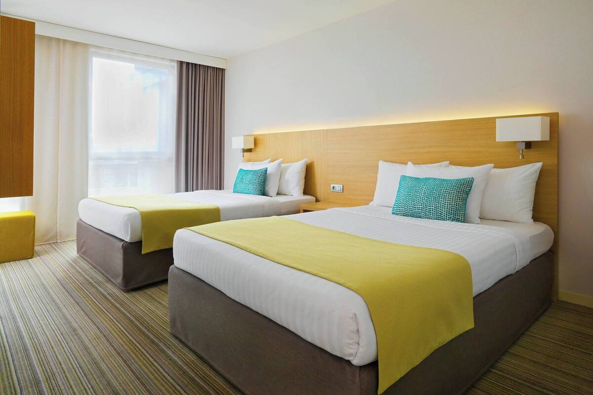 Ein Hotelzimmer mit zwei Betten, jeweils mit Kopfteil, einem Vorhangfenster und einem Teppichboden.