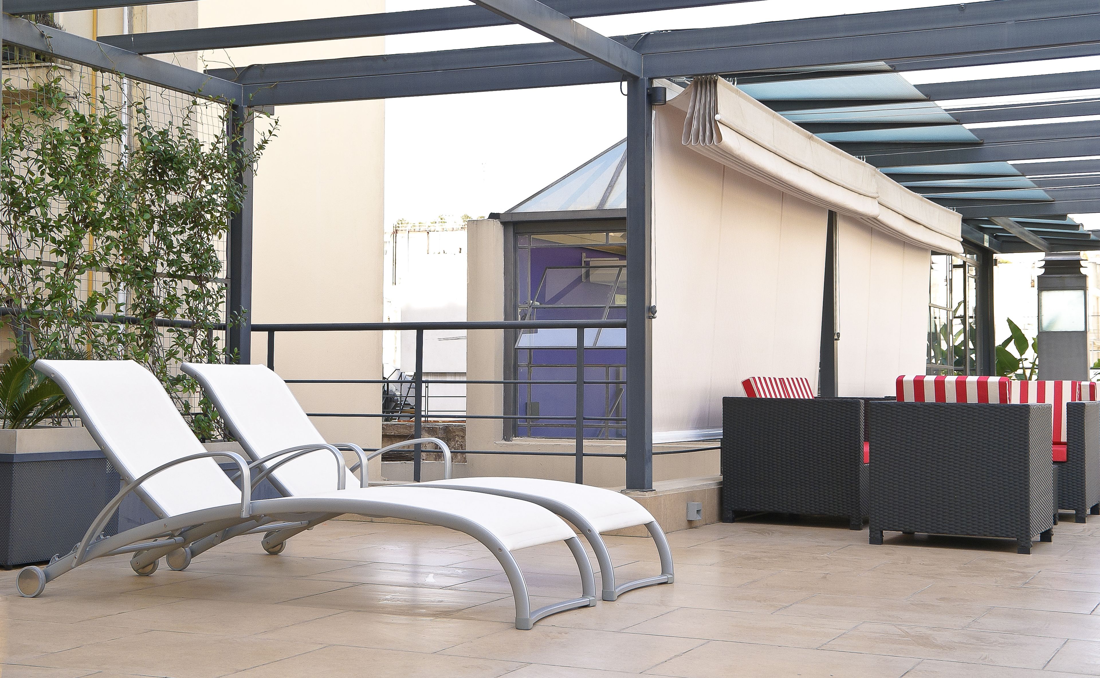 A covered outdoor seating area with lounge chairs and a table, a potted plant, and a building in the background.