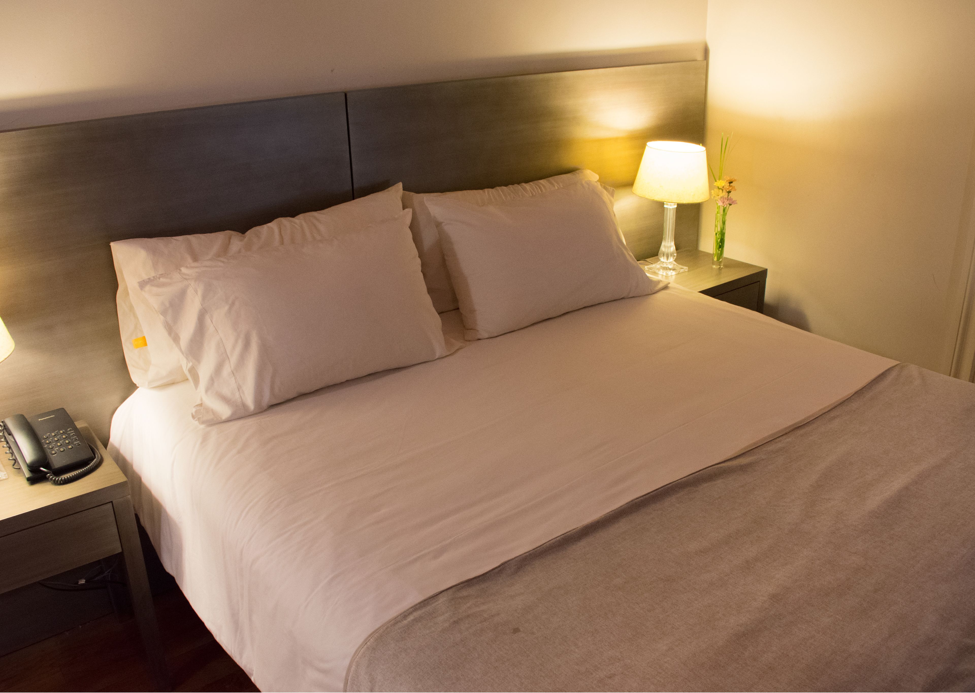 A neatly made bed with white linens and a wooden headboard. There is a bedside table with a lamp and a telephone.