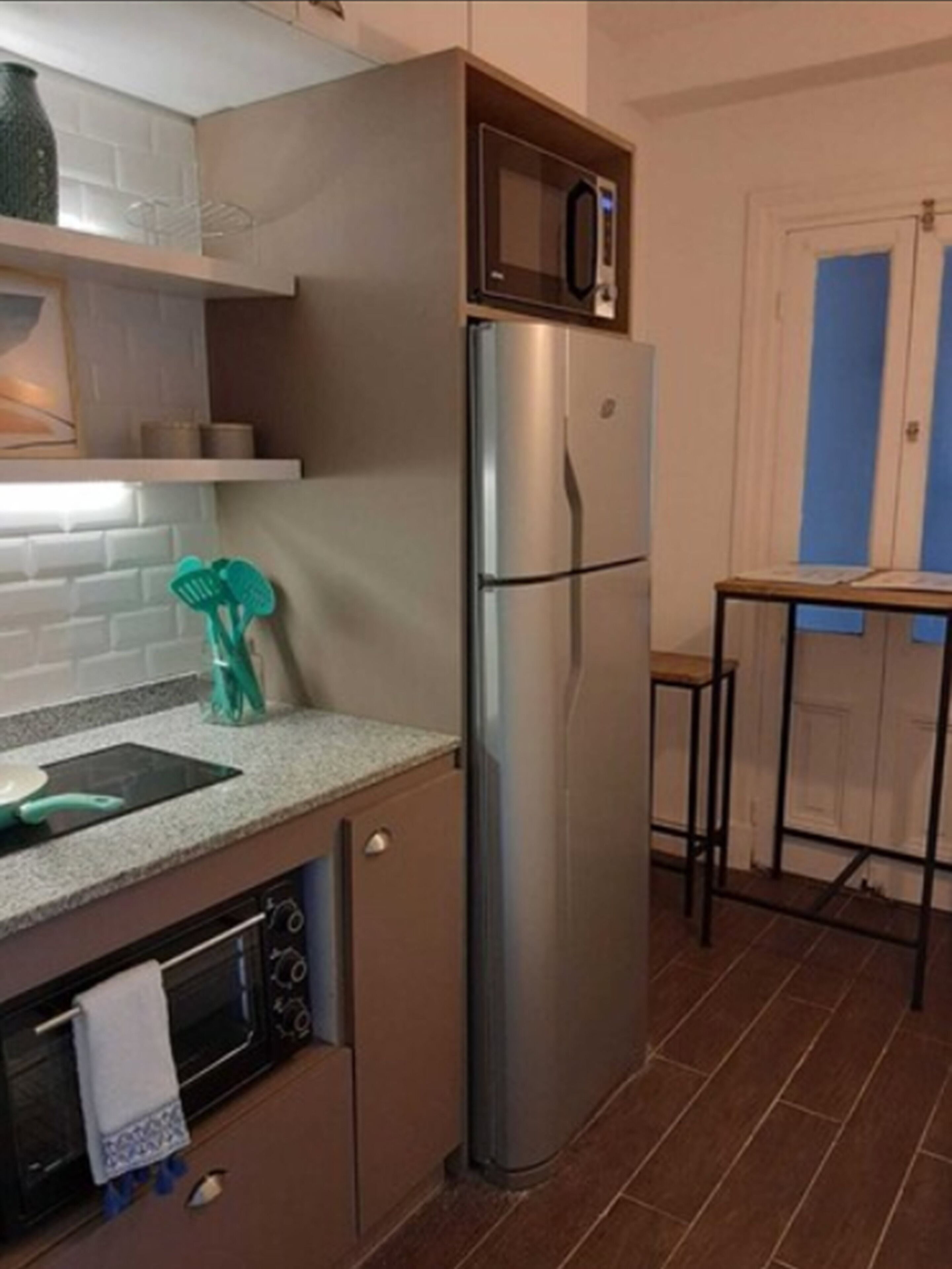 A compact kitchen with a stainless steel refrigerator, a built-in oven, and a microwave. There is a small countertop with a sink and a green kitchen tool.