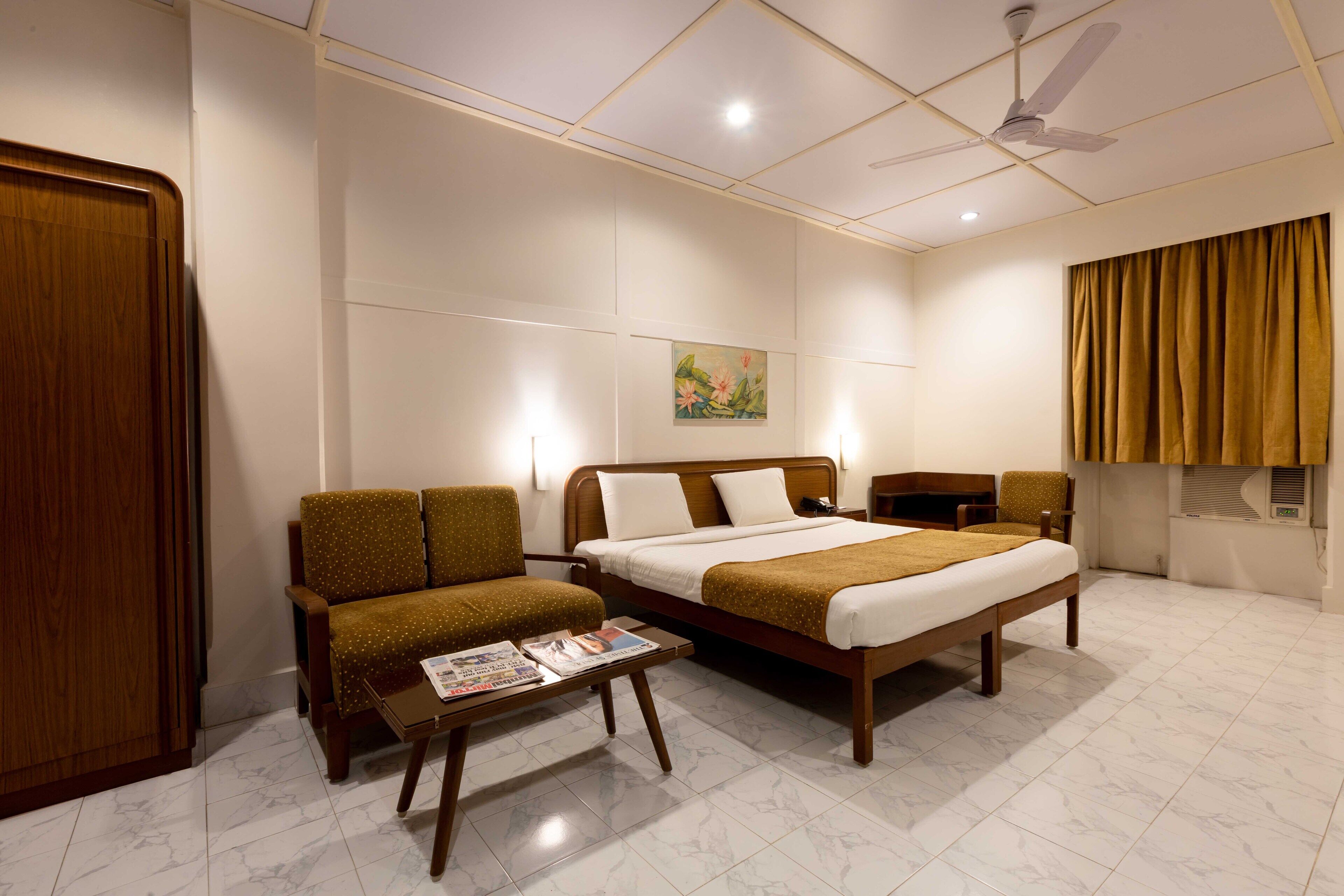 A hotel room with a bed, two armchairs, a small table with a magazine, a ceiling fan, and a painting on the wall.