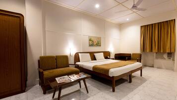 A hotel room with a bed, two armchairs, a small table with a magazine, a ceiling fan, and a painting on the wall.