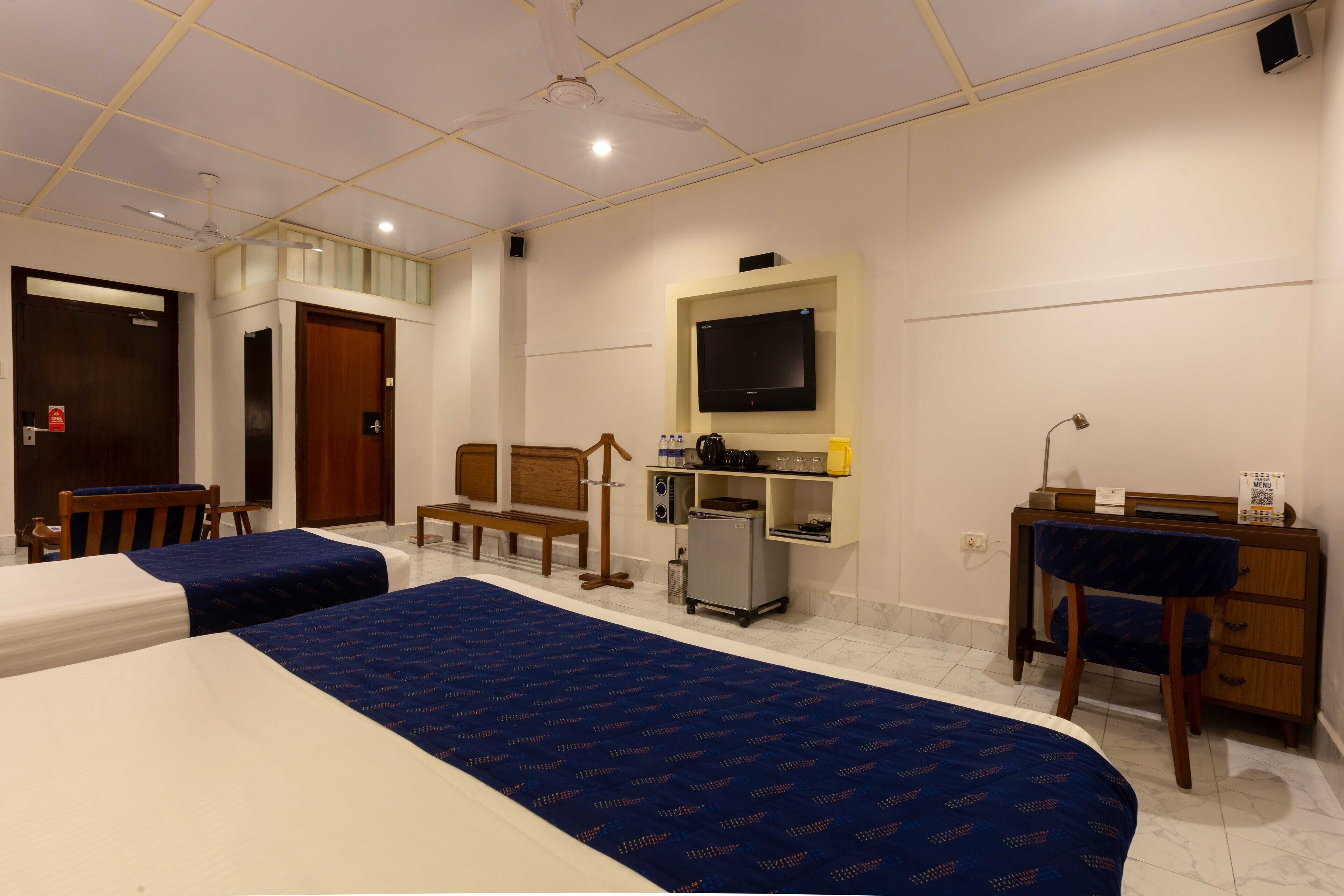 A hotel room with two beds, a desk, a chair, a television, a minibar, and a ceiling fan.