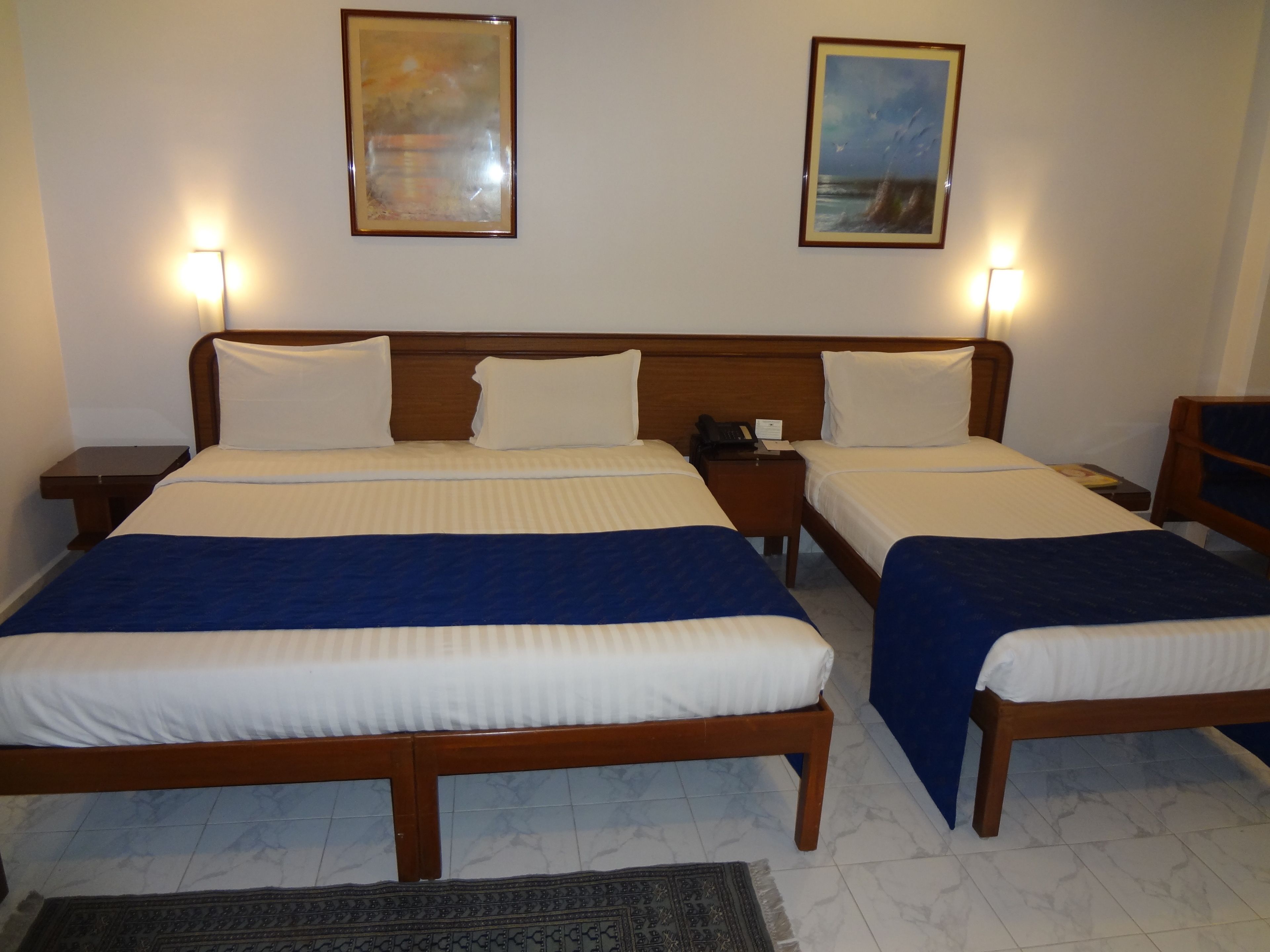 Una habitación de hotel con dos camas, cabecera de madera, mesitas de noche, un velador y dos cuadros en la pared.