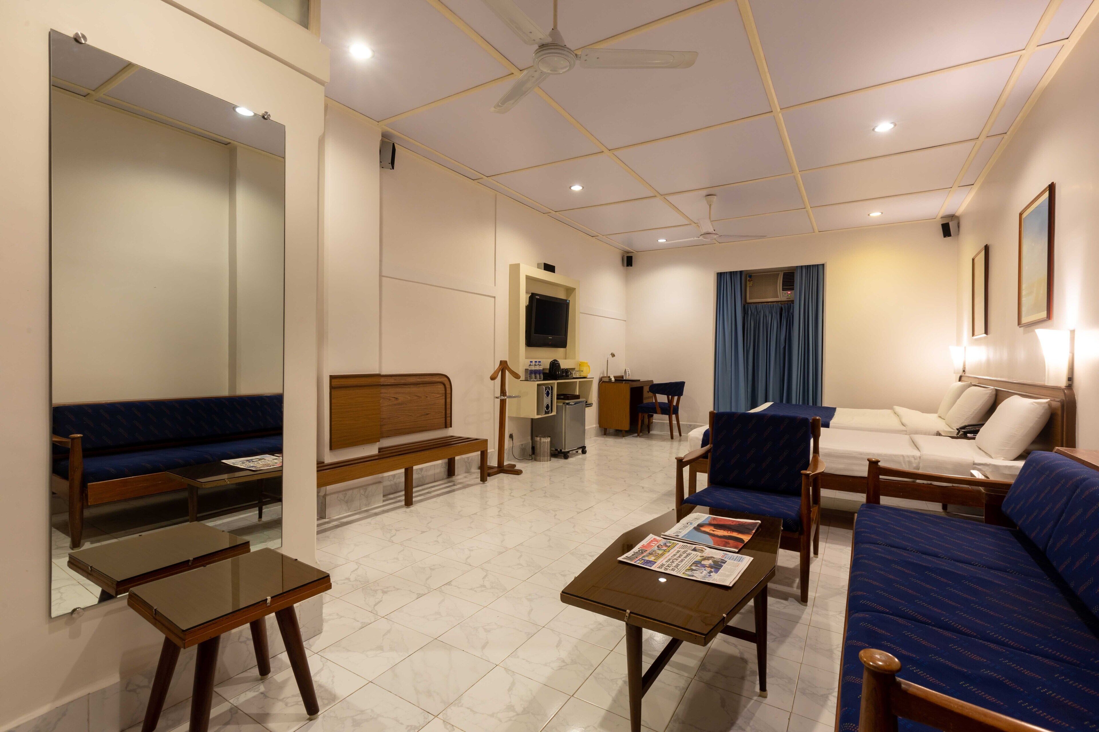 A hotel room with a sofa, a chair, a small table, a mirror, a television, and a desk with a lamp.