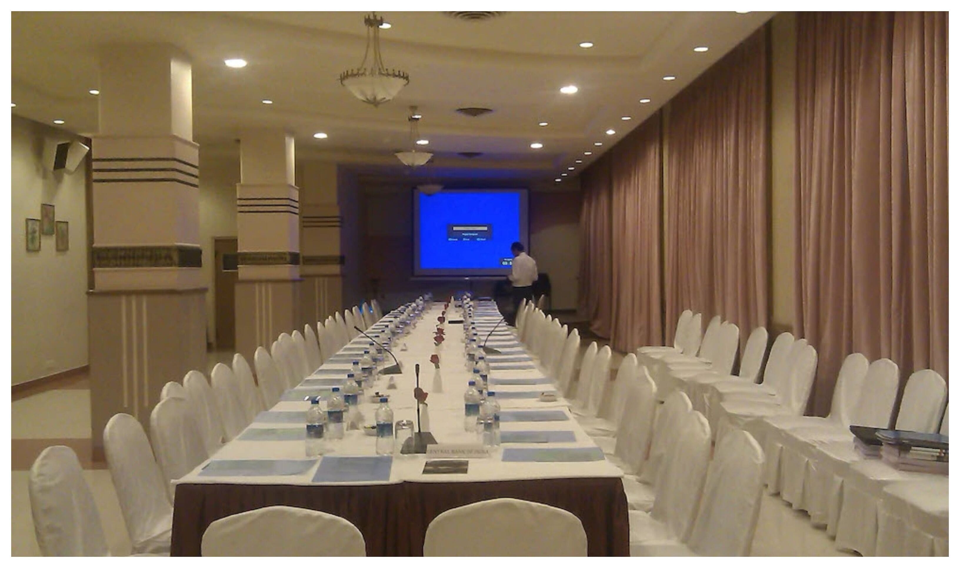 Un salón de conferencias con una larga mesa, sillas blancas y una pantalla para proyector.