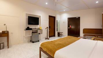 A hotel room with a bed, a TV mounted on the wall, a desk with a chair, and a small kitchenette area.