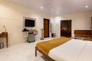 A hotel room with a bed, a TV mounted on the wall, a desk with a chair, and a small kitchenette area.