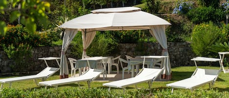 A covered outdoor seating area with white lounge chairs and tables, surrounded by greenery and a stone wall.