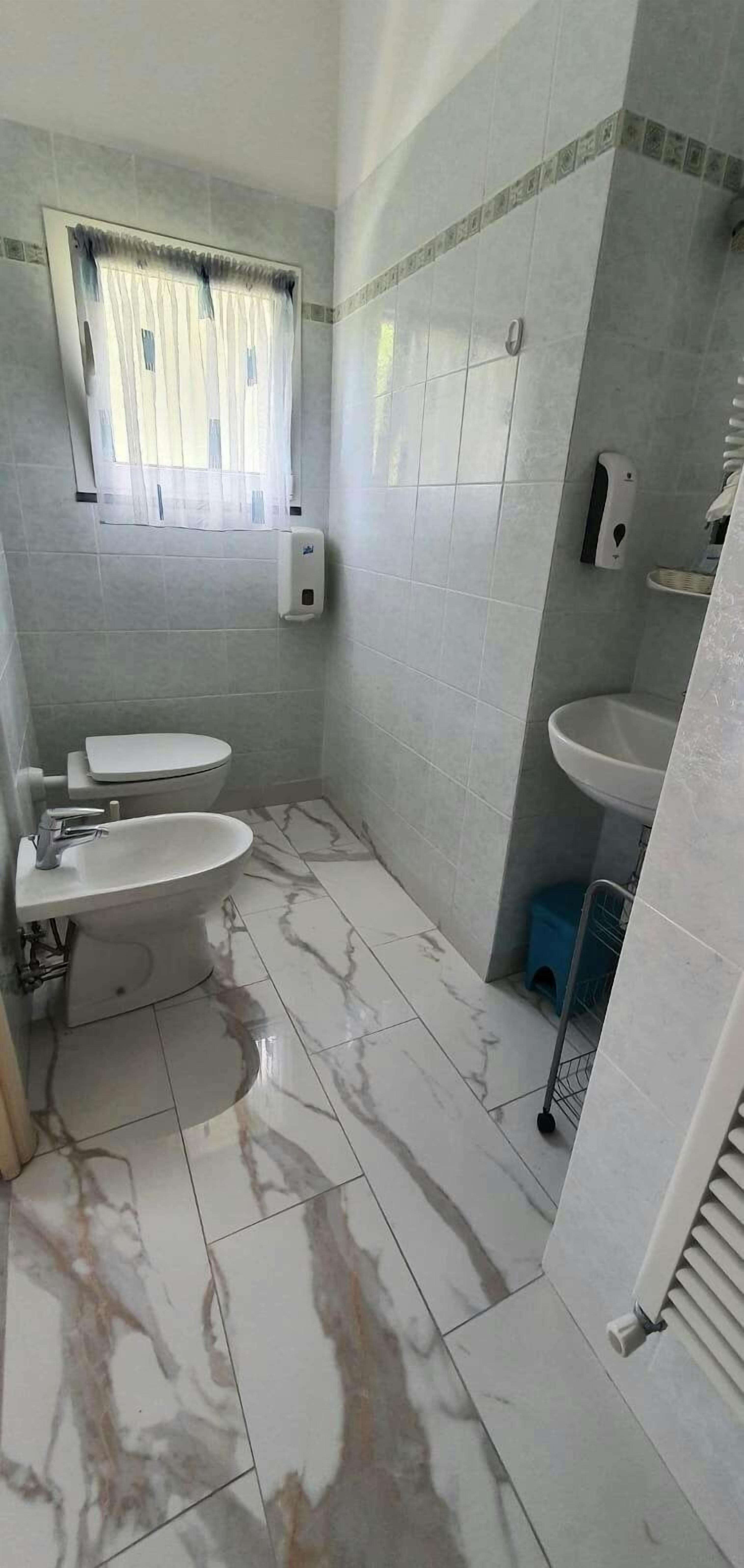 A bathroom with two sinks, a toilet, a window with a curtain, and a hand soap dispenser.