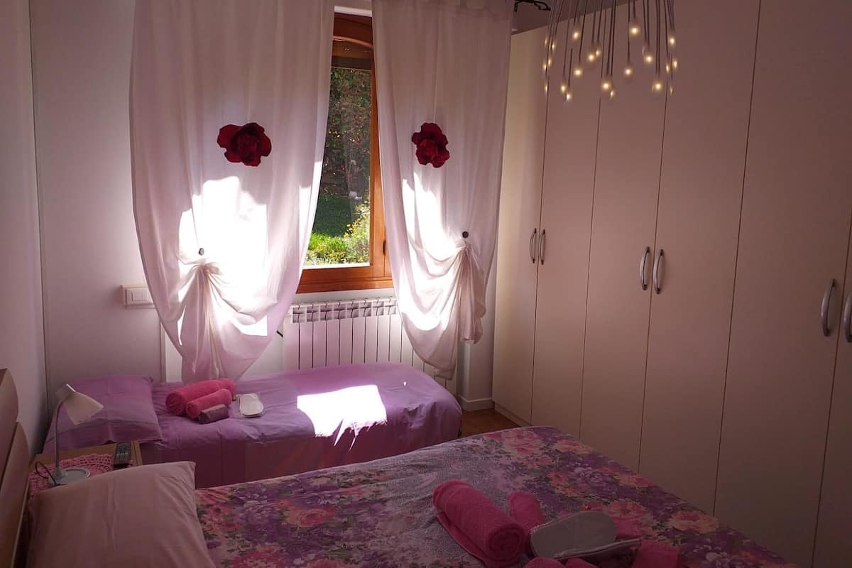 A bedroom with two beds, white curtains, a window with a view of greenery, and a wardrobe.
