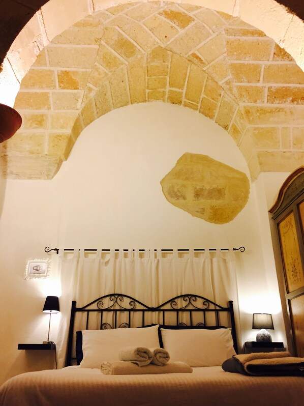 A hotel room with a stone archway, a bed with white linens, two bedside lamps, and a wooden cabinet.