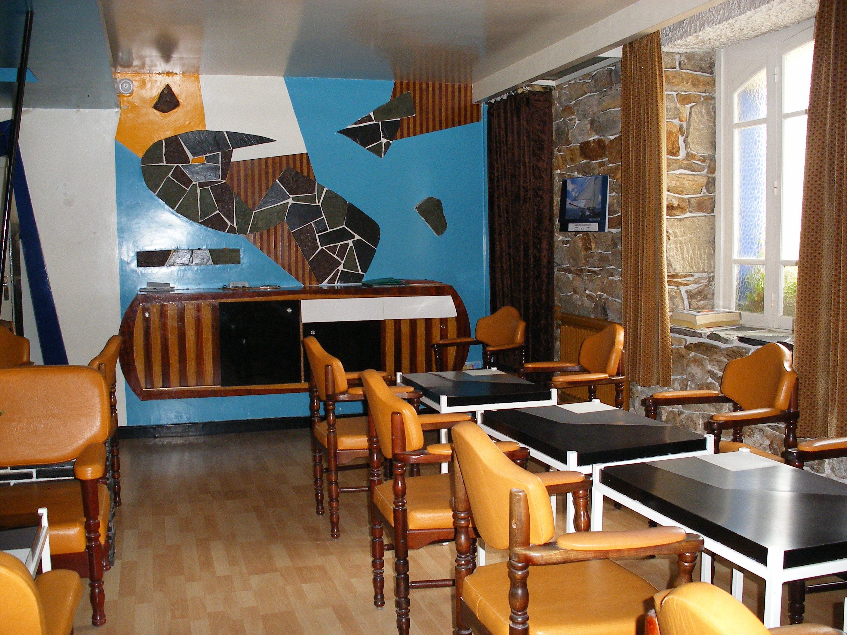 Un restaurante con sillas y mesas de madera, un mural en la pared azul y naranja, y una pared de piedra con una ventana.