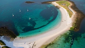 Vista aérea de uma área costeira com águas turquesa cristalinas, uma praia de areia e uma pequena ilha com um edifício.