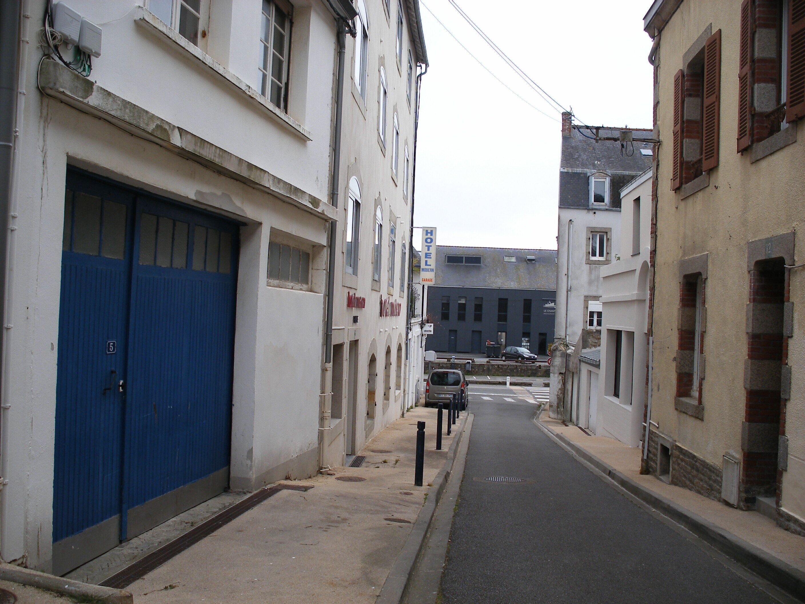 A narrow street with buildings on either side, including a hotel with a blue door and a sign indicating a garage.