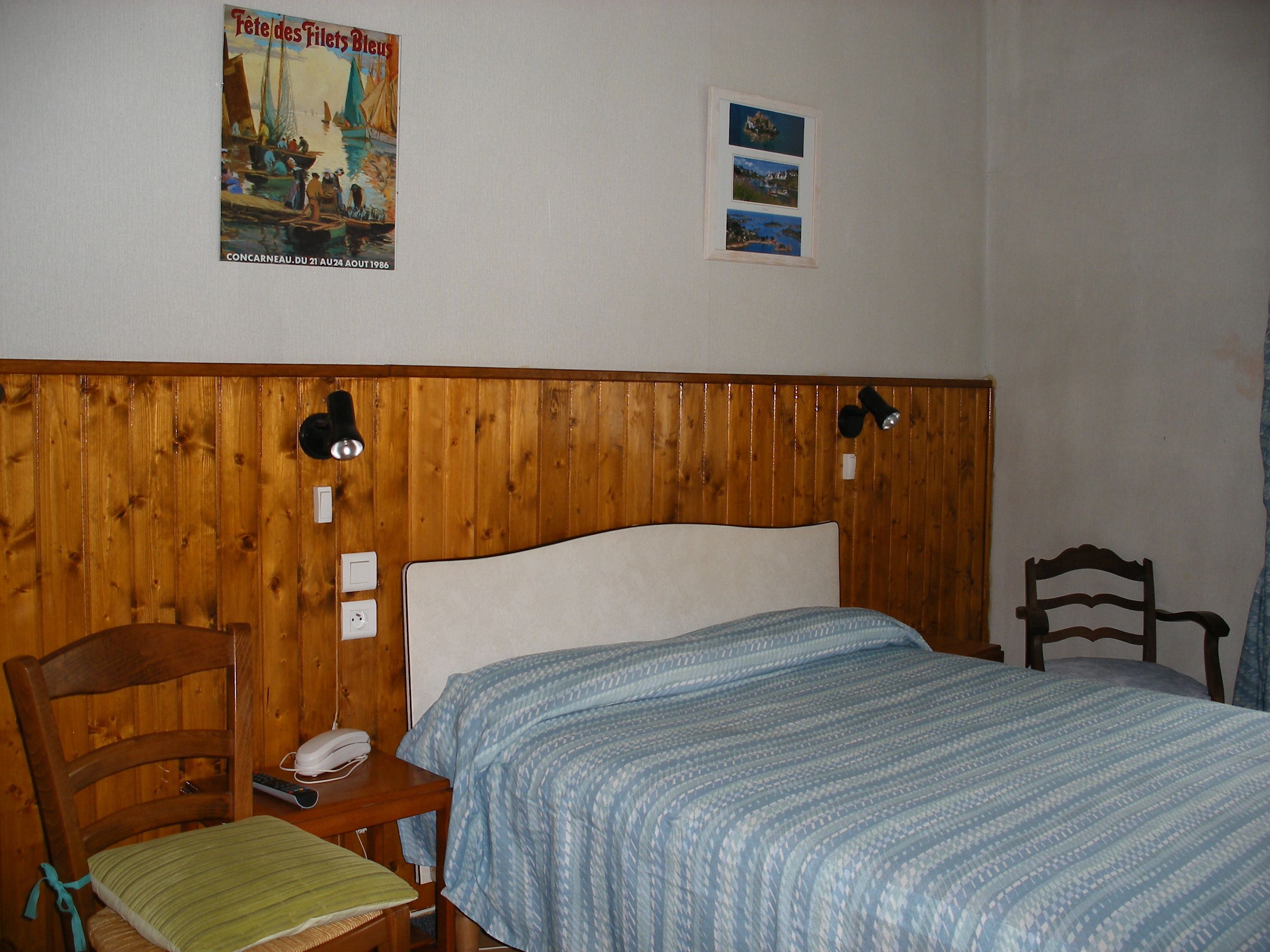 A bedroom with a wooden headboard, a bed with a blue patterned bedspread, a chair with a green cushion, and a poster on the wall.