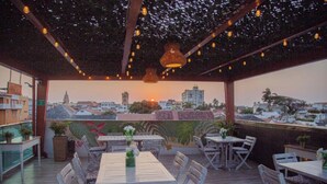 Un patio al aire libre con mesas y sillas, vistas a la ciudad y luces colgantes.