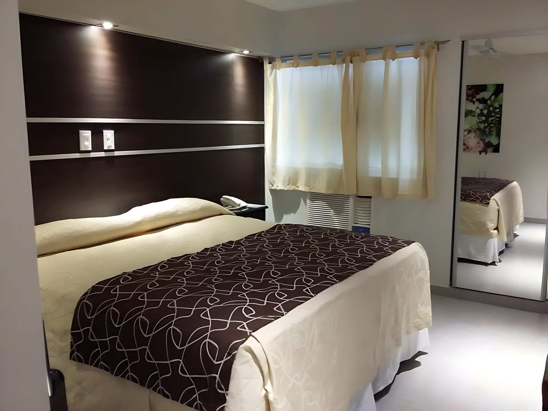 Una habitación de hotel con cama, mesita de noche, espejo y una ventana con cortinas.