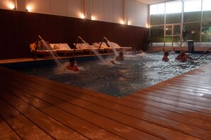 A heated indoor swimming pool with wooden deck, lounge chairs, and a view of the outdoors through large windows. - Platino Termas All Inclusive (Termas de Rio Hondo)